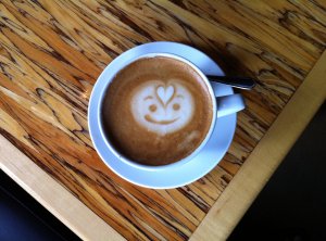 Coffee cup with latte art of a smiling face with a heart on the forehead of the smiling face.