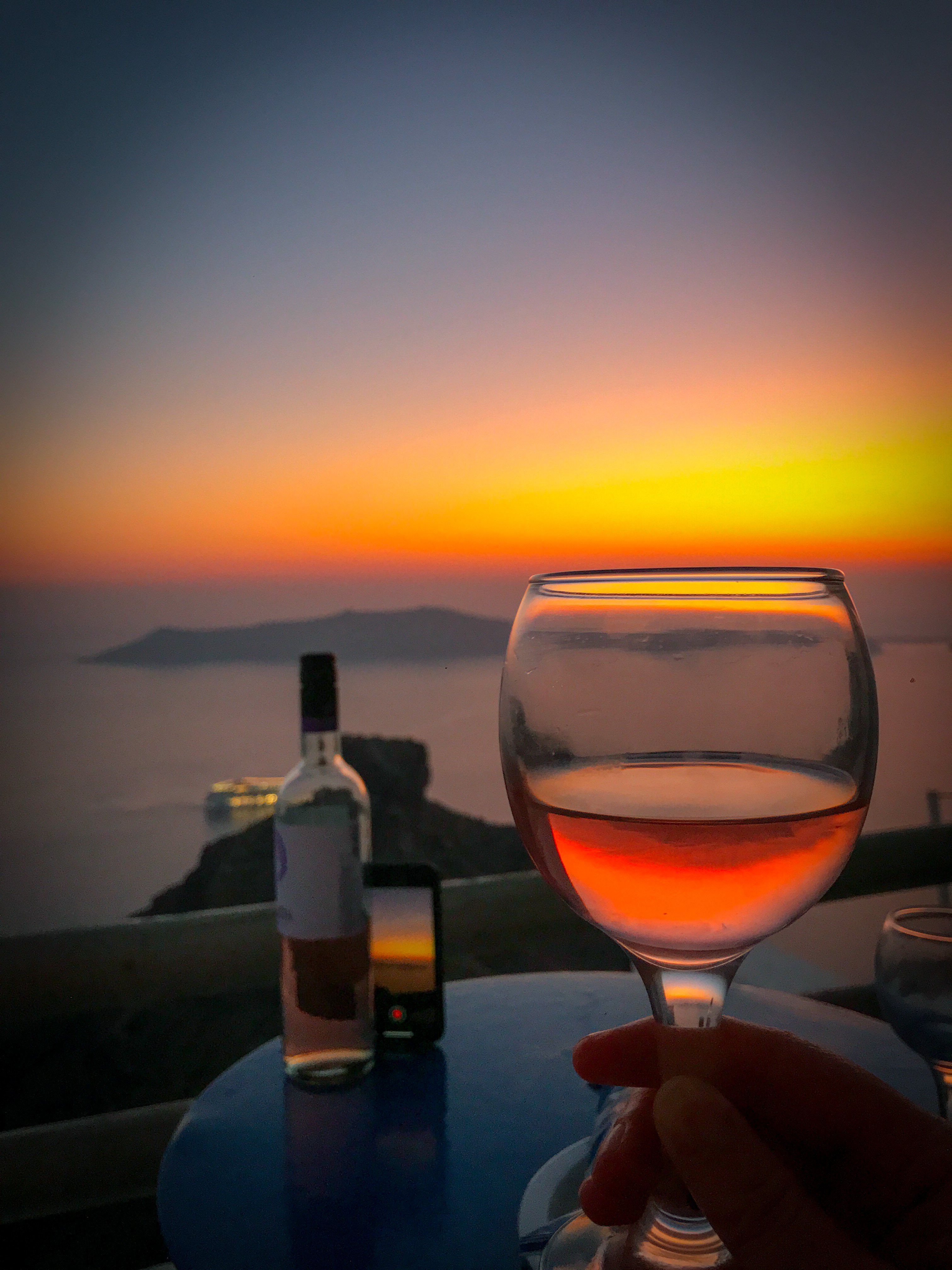 A Santorini Sunset as it reflects through a wineglass and our other phone propped up against the wine bottle as we take a time lapse of the sunset.