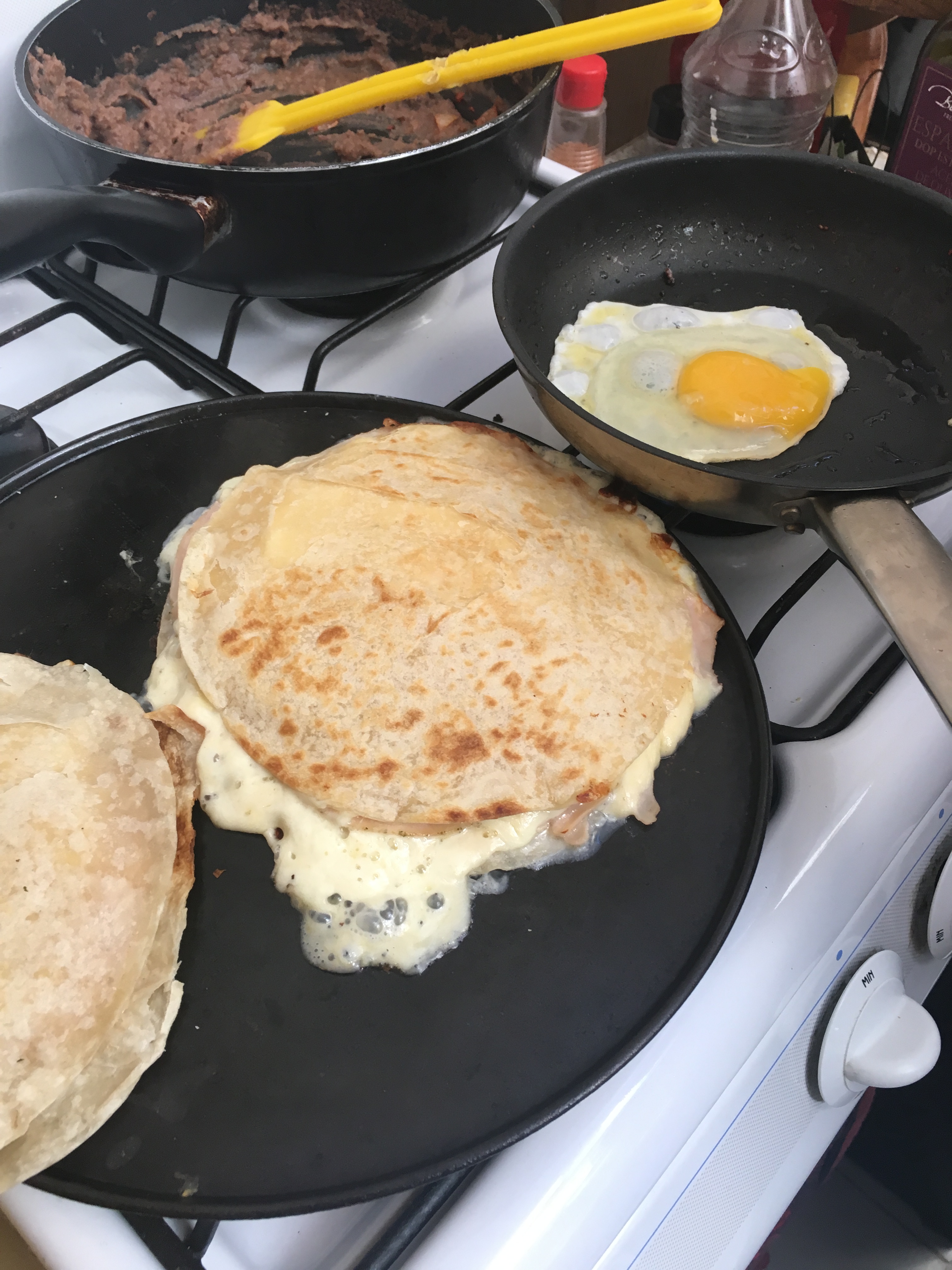 A Mexican breakfast being cooked on the stove with a pot of beans, an egg frying and a quesadilla oozing with melted cheese.